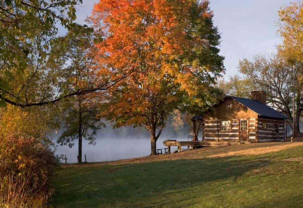 A log cabin sits by a lake surrounded by trees with autumn leaves, where mist rises from the water and sunlight filters through the branches—a serene retreat where travelers seek calm.