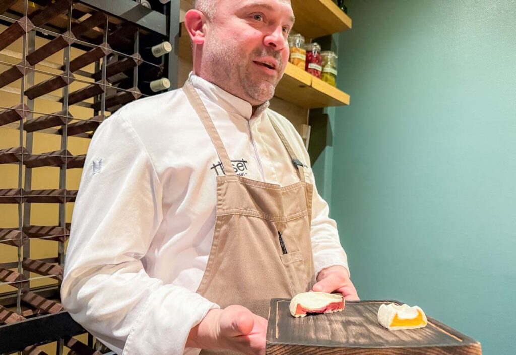 A chef in a white uniform and beige apron holds a wooden tray with two small pieces of food in a kitchen setting.