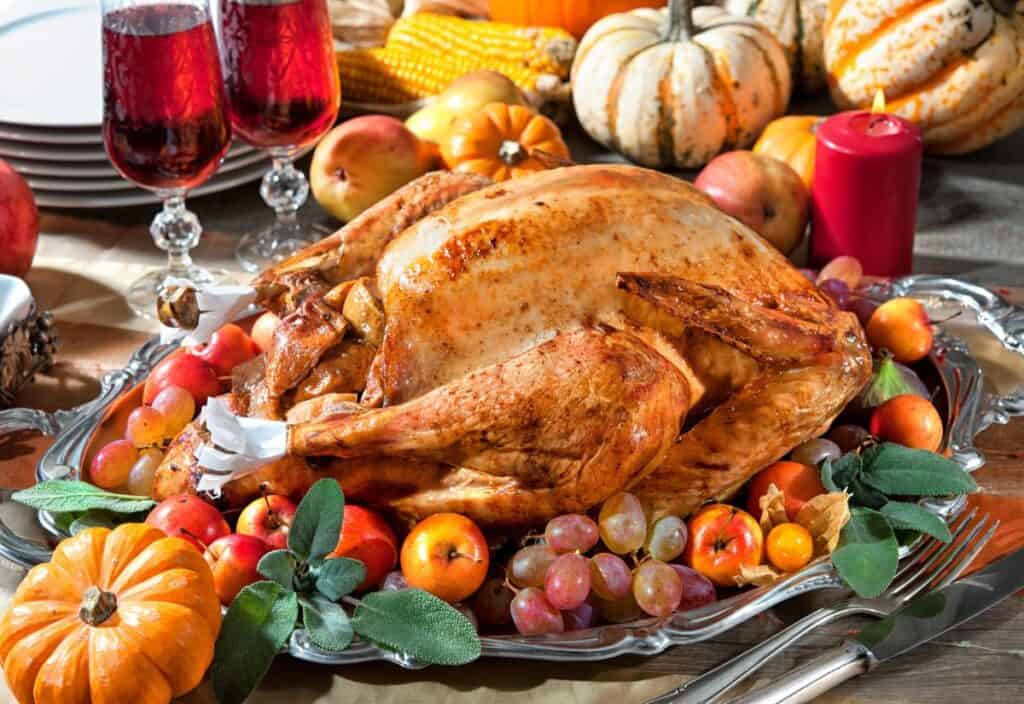 A roasted turkey is served on a platter surrounded by fruits, small pumpkins, and greenery, with wine glasses, candles, and decorative gourds in the background.