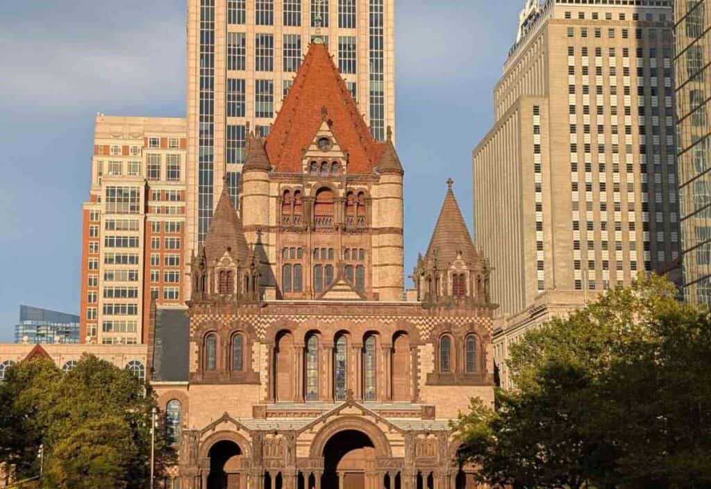 A historic stone church with a red roof stands in front of modern high-rise buildings under a clear sky, surrounded by trees.