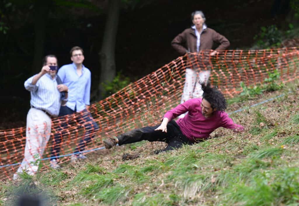 A person in a magenta shirt slides down a grassy, sloped hill near orange netting as three onlookers watch and take photos—capturing the excitement of one of the world’s strangest contests.