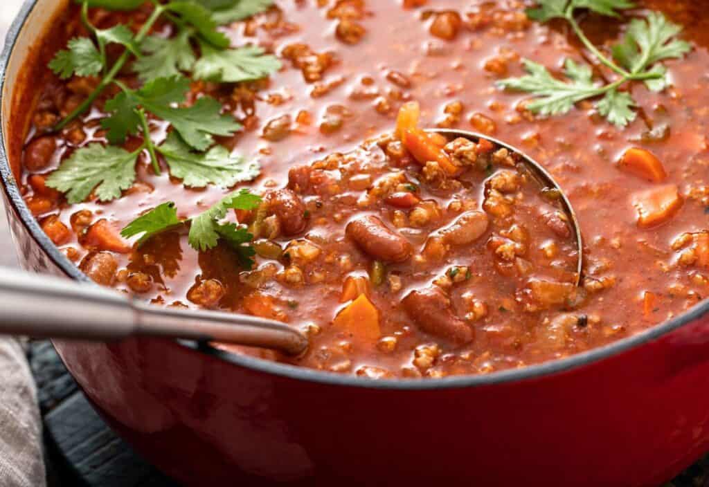 Red pot filled with chili containing ground meat, kidney beans, tomatoes, and carrots, garnished with fresh cilantro, with a ladle resting inside.