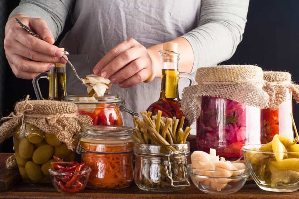 Person arranging assorted jars of pickled vegetables, including olives, peppers, green beans, and cabbage, on a wooden table.