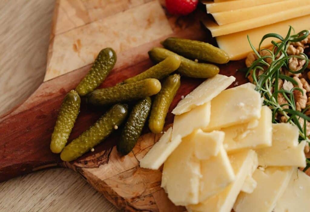 Sliced cheese, pickles, walnuts, and herbs arranged on a wooden serving board.