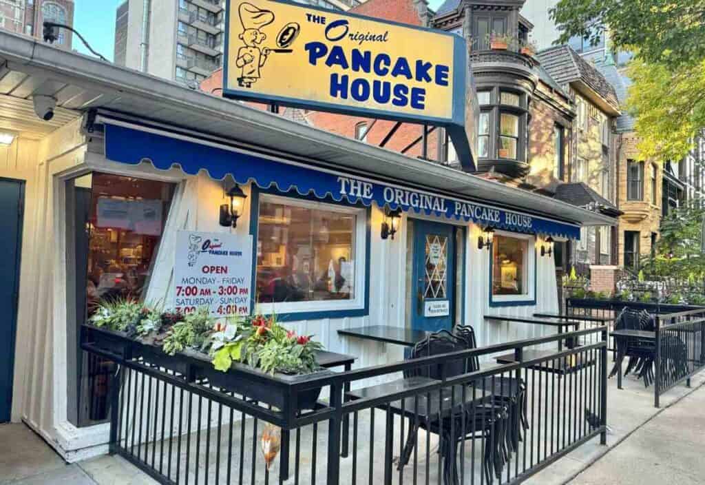 Street view of The Original Pancake House restaurant with blue awning, outdoor seating, and business hours sign by the entrance.