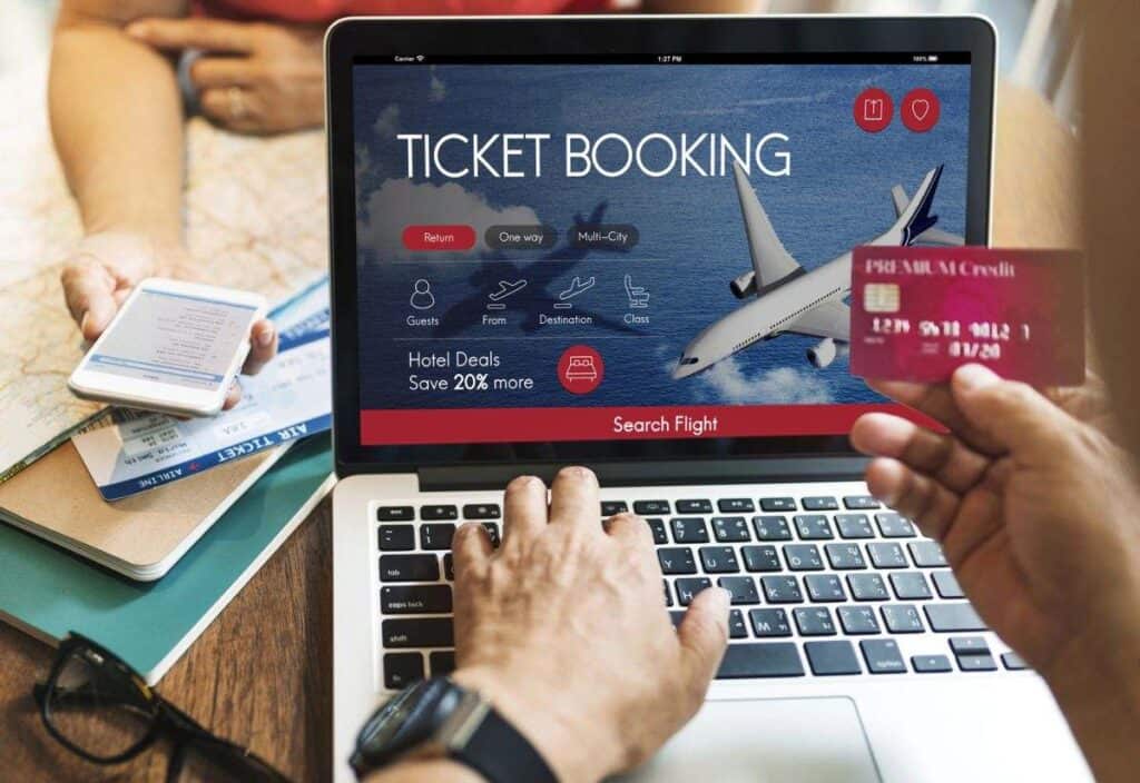 Person booking a flight online on a laptop during Travel Tuesday; one hand types, another holds a credit card, while a second person checks deals on a smartphone.