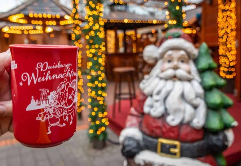 A hand holds a red mug with German writing in front of a Santa Claus statue and festive lights at a Christmas market.